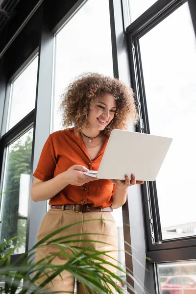 Низкий угол зрения счастливая деловая женщина с вьющимися волосами глядя на ноутбук, стоя возле окон в офисе — стоковое фото