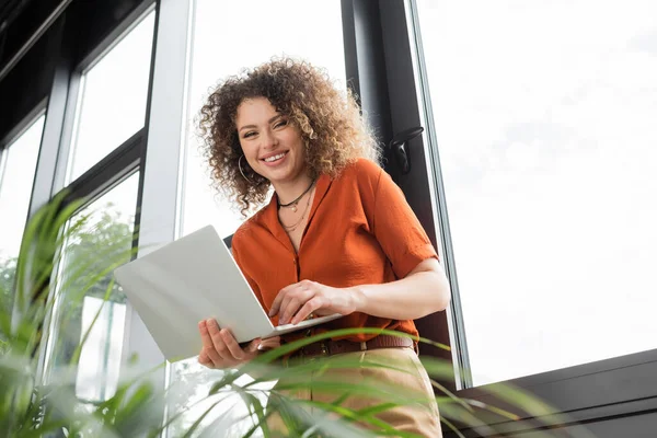Vue à angle bas de femme d'affaires avec les cheveux bouclés tenant ordinateur portable et regardant la caméra à côté de plantes vertes dans le bureau — Photo de stock