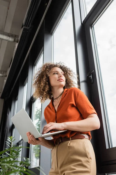 Low angle view of businesswoman with curly hair holding laptop and looking through window in modern office — Stock Photo