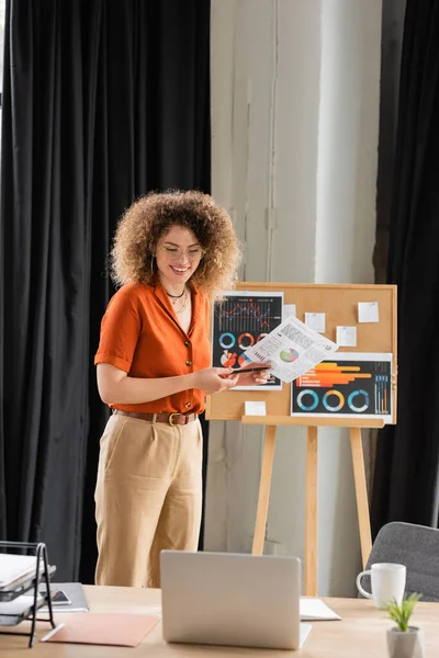 Счастливая деловая женщина в очках, держащая инфографику возле офисной доски во время видеозвонка — стоковое фото