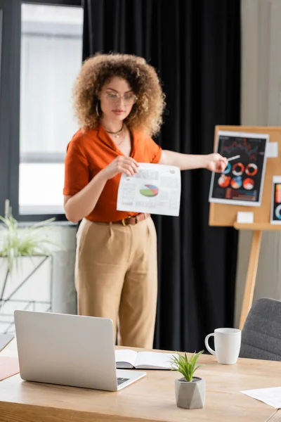Curly businesswoman in glasses pointing with pen at infographics on office board during video call — Stock Photo