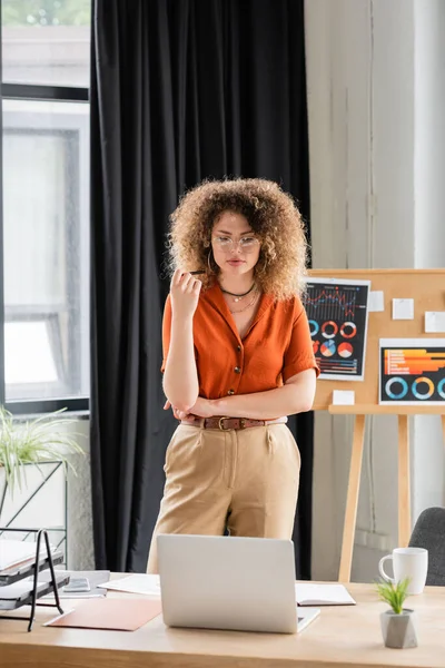 Curly businesswoman in glasses looking at laptop near office board with infographics — Stock Photo
