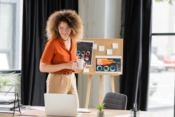 Empresária alegre em óculos segurando copo e olhando para laptop perto de placa de escritório com infográficos — Fotografia de Stock