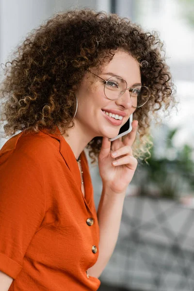 Lockige und glückliche Geschäftsfrau mit Brille, die im Büro auf dem Smartphone spricht — Stockfoto