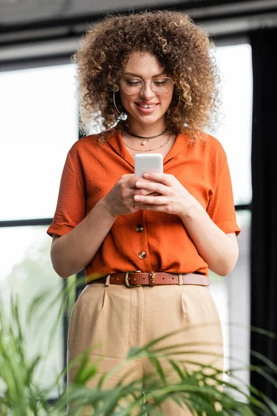 Alegre mujer de negocios en gafas de mensajería en el teléfono inteligente, mientras que de pie en la oficina moderna - foto de stock