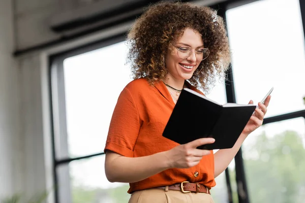 Весела бізнес-леді в окулярах тримає смартфон і блокнот, стоячи в сучасному офісі — стокове фото
