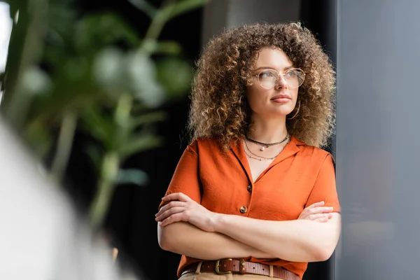 Donna d'affari pensosa con i capelli ricci in piedi con le mani piegate in ufficio moderno — Foto stock