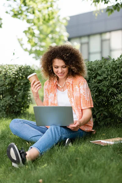 Glückliche Frau mit lockigem Haar hält Pappbecher in der Hand und benutzt Laptop, während sie auf Gras sitzt — Stockfoto