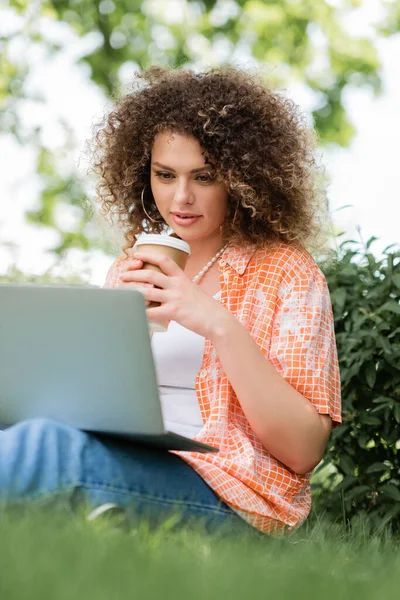 Jeune pigiste avec les cheveux bouclés tenant tasse en papier et en utilisant un ordinateur portable tout en étant assis sur l'herbe — Photo de stock
