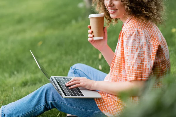 Обрезанный вид веселого фрилансера с вьющимися волосами, держащего бумажную чашку и использующего ноутбук, сидя на траве — стоковое фото