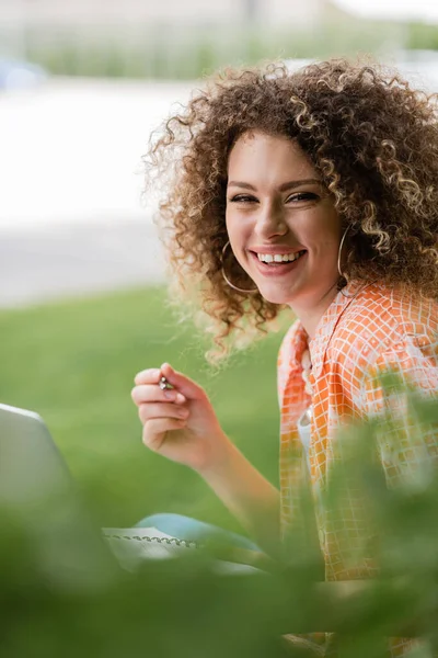 Positive freelancer woman with curly hair using laptop and holding pen in park — Stock Photo
