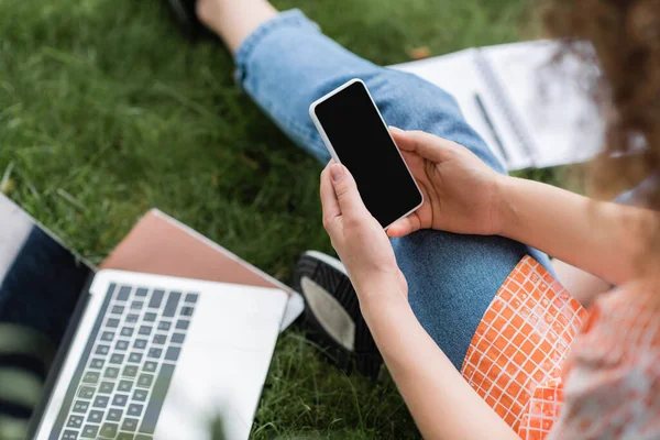 Ausgeschnittene Ansicht einer jungen Frau, die ihr Smartphone mit leerem Bildschirm hält, während sie neben Laptop und Notizbuch im Gras sitzt — Stockfoto
