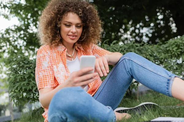 Low angle view of positive freelancer woman with curly hair using smartphone in park — Stock Photo
