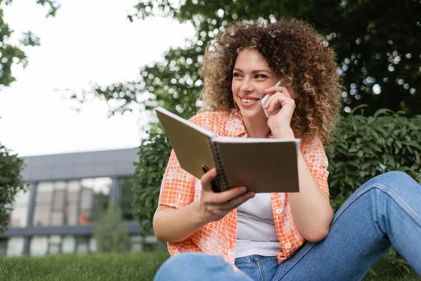 Happy young woman with curly holding notebook while talking on smartphone in green park — Stock Photo