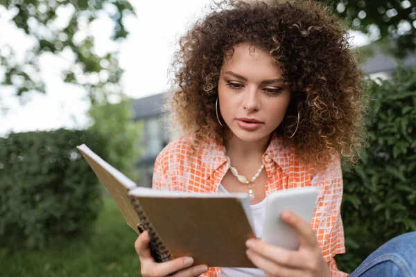 Pretty young woman with curly holding notebook while using smartphone in green park — Stock Photo