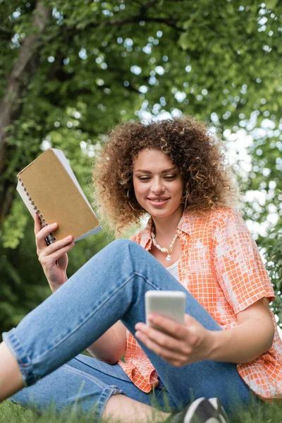 Low angle view of smiling young woman with curly holding notebook while using smartphone in green park — Stock Photo