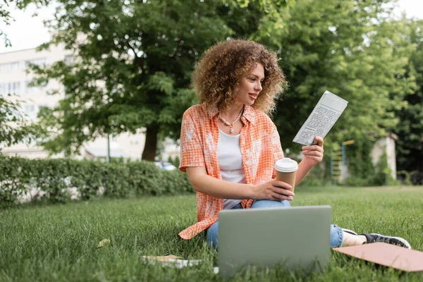 Donna allegra con i capelli ricci leggendo giornale e tenendo il caffè per andare mentre seduto sull'erba vicino al computer portatile — Foto stock