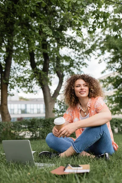 Sorglose Frau hält Pappbecher in der Hand und sitzt auf grünem Gras neben Laptop — Stockfoto