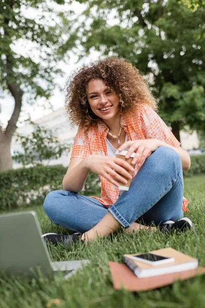 Happy freelancer woman holding paper cup and sitting on green grass near laptop — Stock Photo