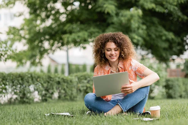 Happy freelancer woman using laptop while sitting on green lawn near paper cup — Stock Photo