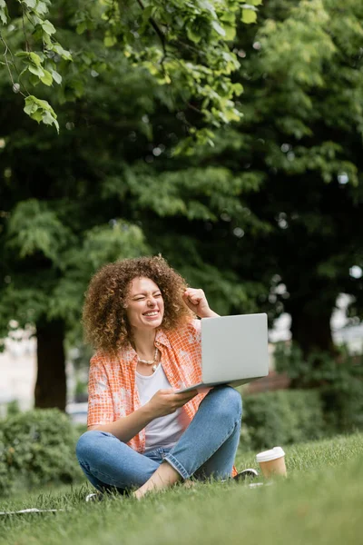 Mujer freelancer emocionada con los ojos cerrados sosteniendo el ordenador portátil y sentado en el césped en el parque verde - foto de stock