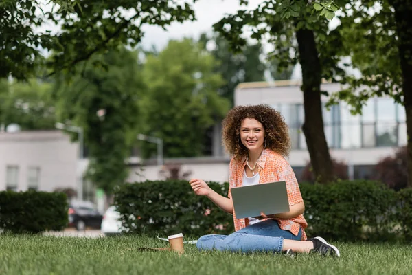 Cheerful freelancer woman with curly hair holding laptop while working remotely in green park — Stock Photo