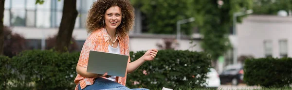 Cheerful freelancer woman with curly hair holding laptop while working remotely in green park, banner — Stock Photo