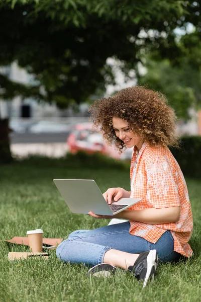 Cheerful freelancer woman with curly hair using laptop while working remotely in green park — Stock Photo