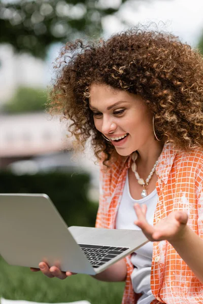 Довольна фрилансер женщина с кудрявыми волосами держа ноутбук во время работы удаленно в зеленом парке — стоковое фото