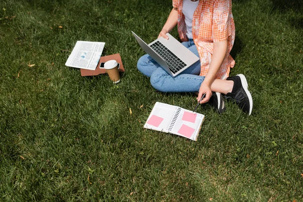 Високий кут зору обрізаної жінки за допомогою ноутбука, сидячи на газоні біля паперової чашки та блокнота — стокове фото