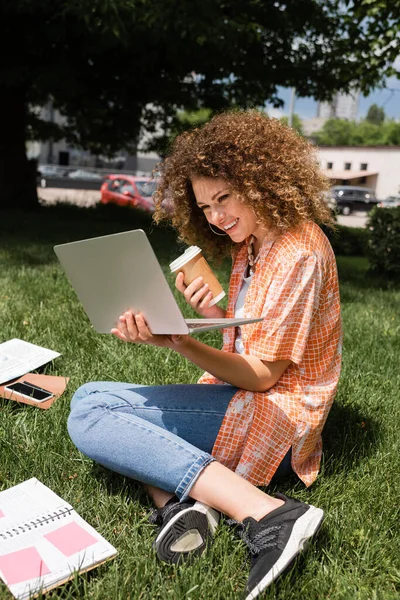 Glückliche und lockige Freiberuflerin, die Pappbecher und Laptop hält, während sie aus der Ferne im Park arbeitet — Stockfoto
