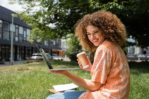 Fröhliche und lockige Freiberuflerin, die Pappbecher und Laptop in der Hand hält, während sie fernab im Park arbeitet — Stockfoto