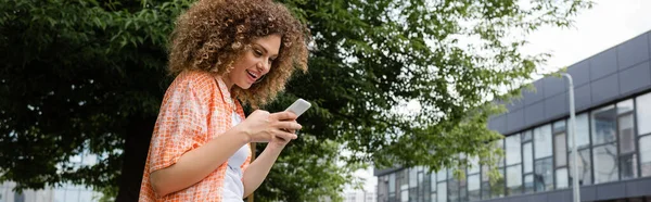 Cheerful woman with curly hair messaging on smartphone in green park, banner — Stock Photo