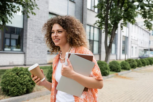 Glückliche Frau mit lockigem Haar mit Coffee to go und Laptop mit Ordner, während sie draußen spaziert — Stockfoto