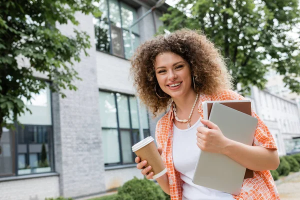 Overjoyed woman with curly hair holding coffee to go and laptop with folder while walking outside — Stock Photo