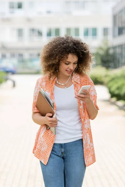Pleased woman with curly hair using smartphone and holding laptop with folder while walking outside — Stock Photo