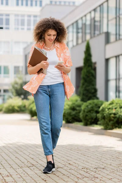 Full length of cheerful woman with curly hair using smartphone and holding laptop with folder while walking outside — Stock Photo