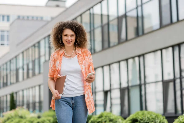 Cheerful woman with curly hair holding smartphone and folder while walking outside — Stock Photo