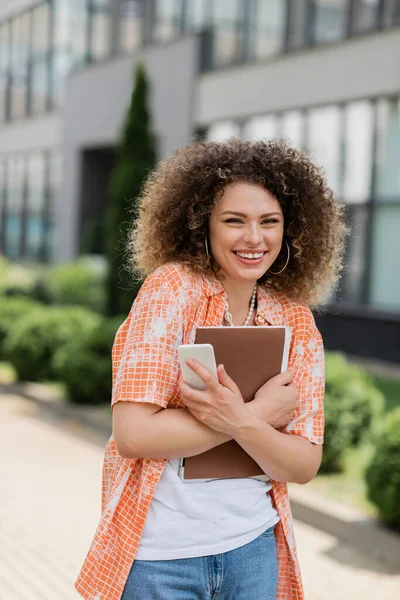 Cheerful woman with curly hair holding smartphone and folder while smiling outside — Stock Photo