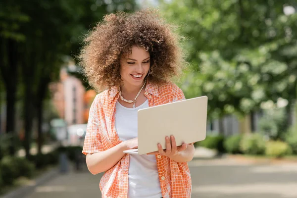 Femme freelance bouclée souriant tout en tenant ordinateur portable et travaillant à distance dans le parc — Photo de stock