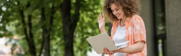 Donna allegra sorridente mentre sventola la mano durante la videochiamata sul computer portatile nel parco verde, banner — Foto stock