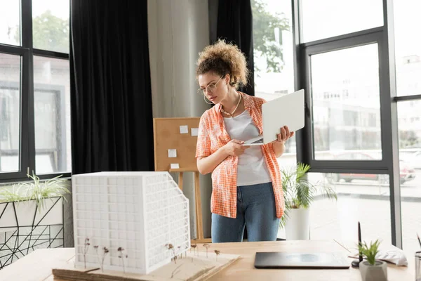 Кучерявий архітектурний дизайнер тримає ноутбук, дивлячись на модель будинку в офісі — стокове фото