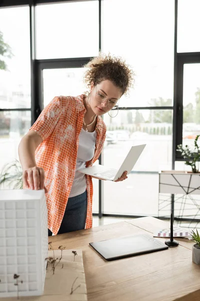 Curly architectural designer holding laptop while checking house model in office — Stock Photo