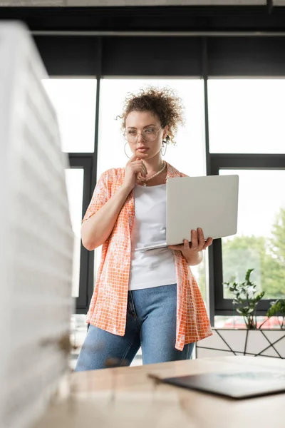Designer de arquitetura encaracolado segurando laptop enquanto olha para o modelo de casa borrada no escritório — Fotografia de Stock