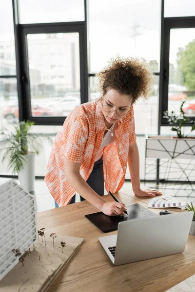 Curly designer using graphic tablet while looking at house model near laptop in office — Stock Photo