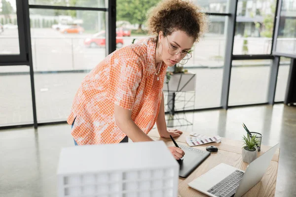 Joven diseñador en gafas usando tableta gráfica mientras mira el modelo de casa cerca de la computadora portátil en la oficina - foto de stock