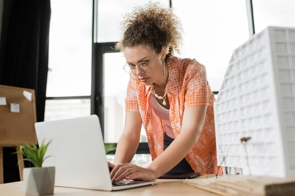 Curly architectural designer using laptop near residential house model on desk — Stock Photo