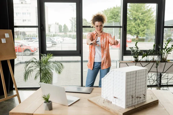 Junge Designerin macht Geste mit den Händen, während sie das Wohnhausmodell auf dem Schreibtisch betrachtet — Stockfoto