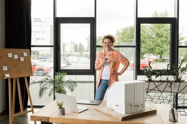 Designer de arquitetura feliz de pé com a mão no quadril perto de gadgets e modelo de casa residencial na mesa — Fotografia de Stock