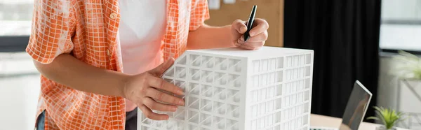 Обрізаний вид архітектурного дизайнера, що тримає ручку стилуса біля моделі житлового будинку в офісі, банер — стокове фото